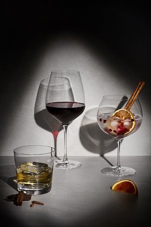 Three stemmed and rocks glasses filled with red wine, clear cocktail, and whiskey with ice, arranged on a table against a dark background.