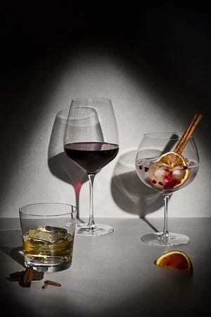 Three stemmed and rocks glasses filled with red wine, clear cocktail, and whiskey with ice, arranged on a table against a dark background.