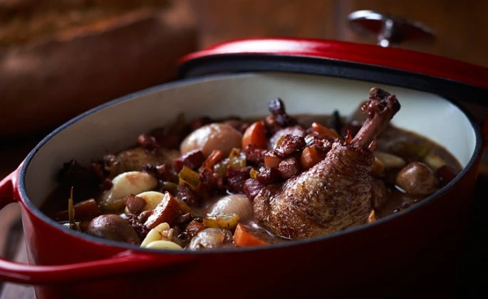 Duck confit in a red cast iron pot with small vegetables, set against a dark background. Close-up shot focusing on the cooked food.
