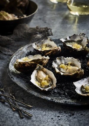 Overhead shot of oysters on a dark plate with forks, featuring fresh seafood delicacies, served alongside wine and bread.