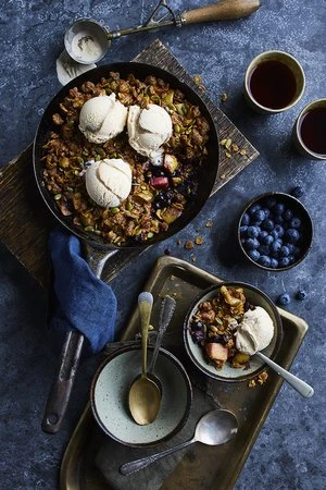 Overhead shot of apple crumble with ice cream in a skillet, served with coffee and blueberries on a rustic surface.