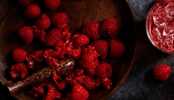 Overhead shot of fresh raspberries on a wooden plate with a muddler, beside a glass of red liquid with ice cubes on a dark surface.