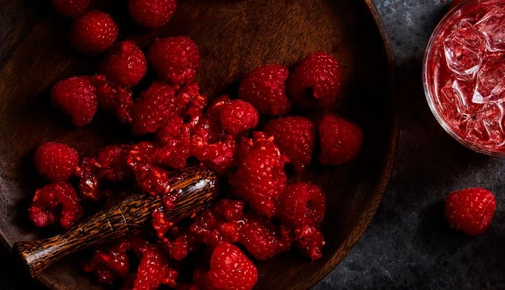 Overhead shot of fresh raspberries on a wooden plate with a muddler, beside a glass of red liquid with ice cubes on a dark surface.