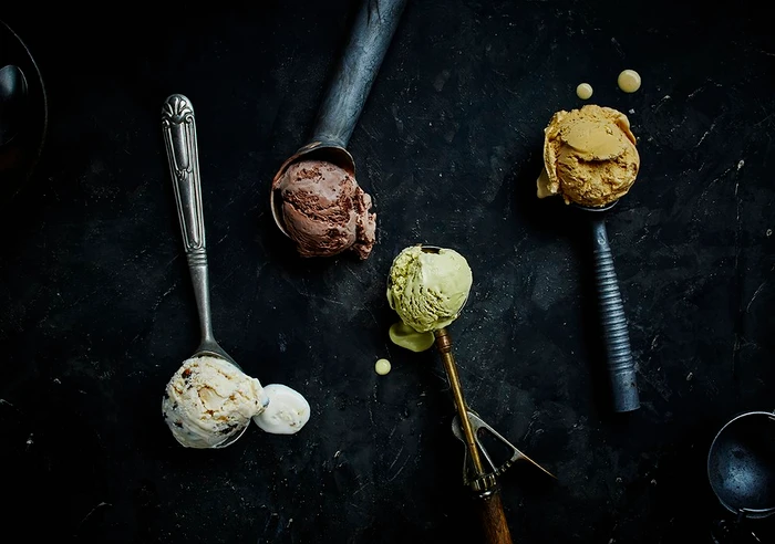 Overhead food photography of four scoops of ice cream with metal scoops on a dark textured surface by Mike Long