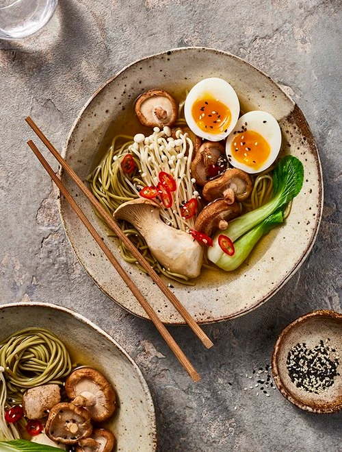 Bowl of noodle soup with sliced mushrooms, halved soft-boiled eggs, and chopsticks on a textured grey surface