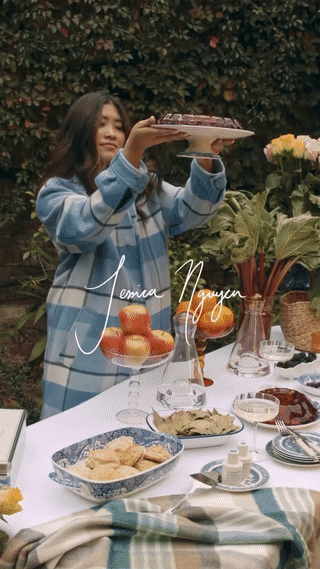 Young Asian woman in blue plaid coat holding cake outdoors. Table with fruit, cookies, drinks, and flowers. Natural light, soft focus.