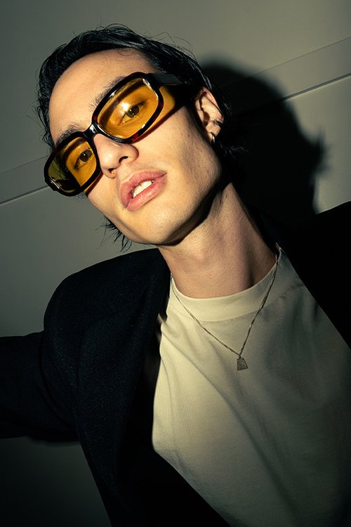 Portrait of a young adult male wearing yellow sunglasses and a black jacket, posing indoors with a serious expression.