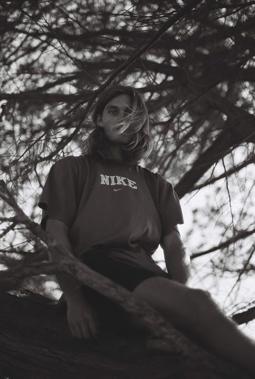 Young adult male in a Nike t-shirt sits on a tree branch outdoors, framed by tree branches in a black and white portrait.