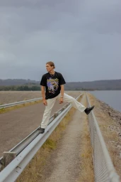 Young man in a black graphic t-shirt and white pants stepping over a metal guardrail near a large body of water under an overcast sky