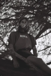 Monochrome portrait of a long-haired man wearing a Nike t-shirt seated in a tree branch against a bright sky.