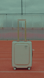 A beige wheeled suitcase standing upright on a red running track with white lane markings under an overcast sky.