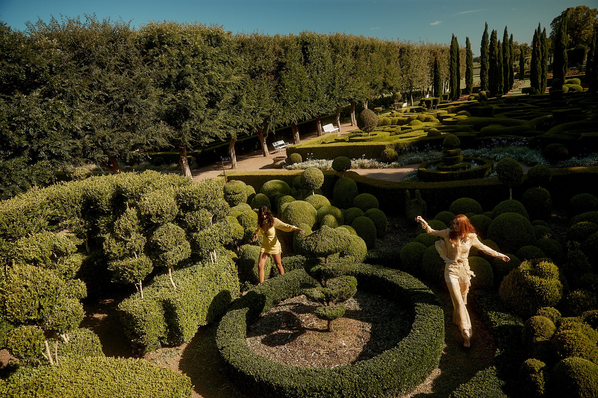 Two women in light-colored outfits stand among intricately designed green topiary in a sunny garden under a clear blue sky.