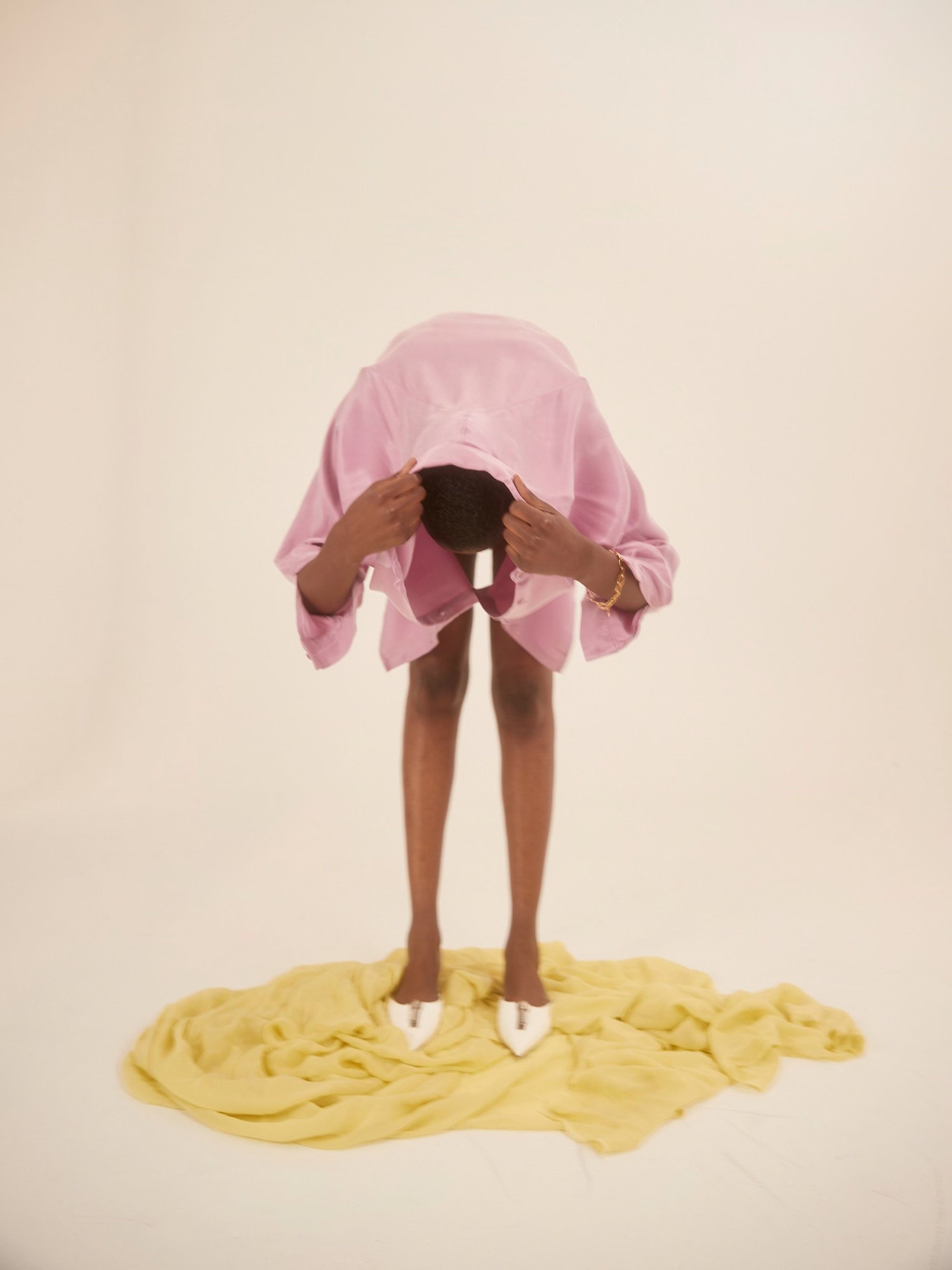 Adult with medium skin tone bending forward, wearing a pink shirt and white high heels, standing on a yellow fabric against a white backdrop.