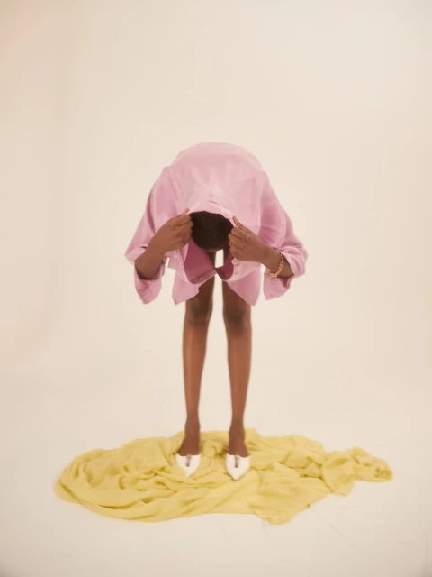 Adult with medium skin tone bending forward, wearing a pink shirt and white high heels, standing on a yellow fabric against a white backdrop.