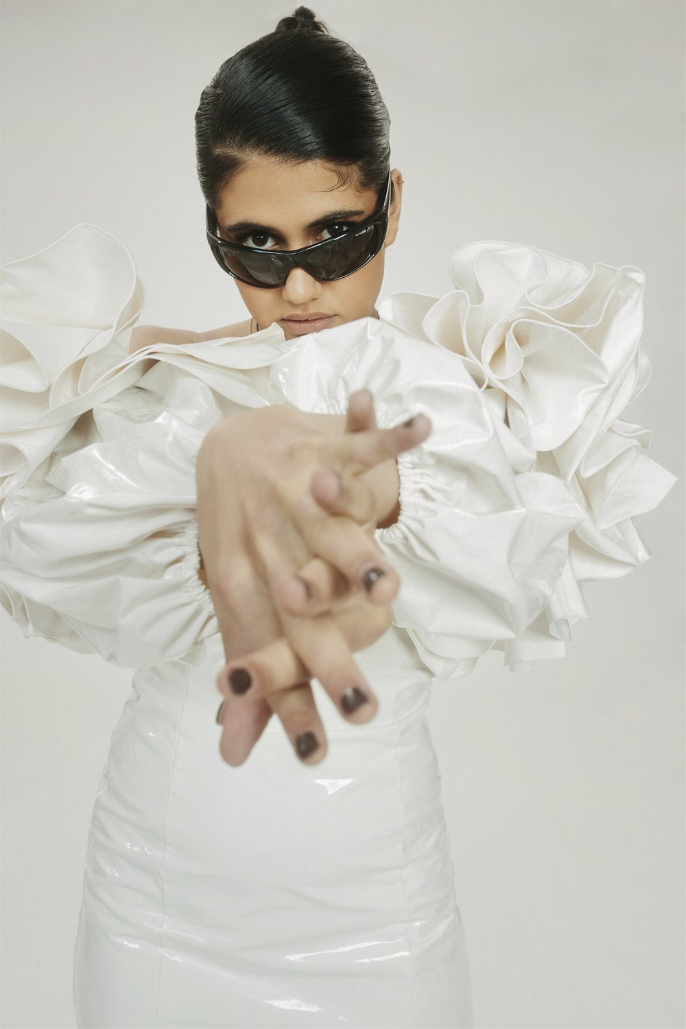 Fashionable woman in white dress and sunglasses poses with hands outstretched in a bright photo studio setting. Editorial fashion shot.