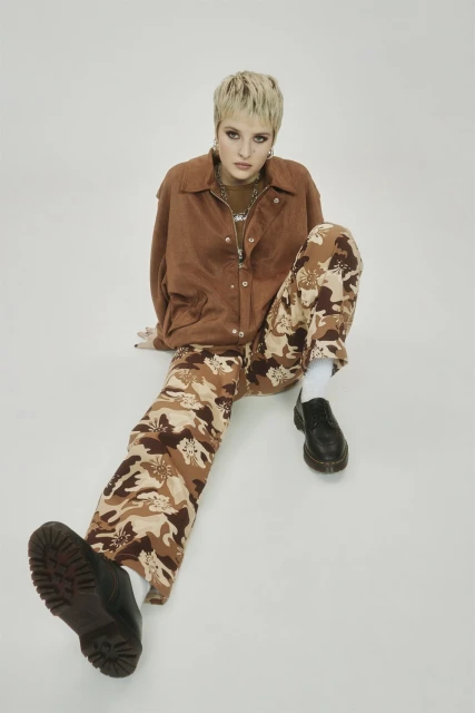 A woman with short blonde hair, wearing a brown corduroy jacket and camouflage pants, sits in a photo studio against a white backdrop.