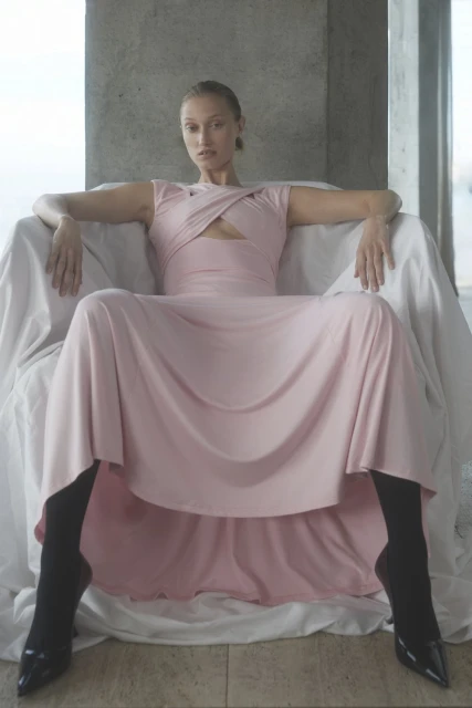 A slender woman in a pink dress sits in a chair draped with white fabric in front of concrete pillars, fashion photoshoot.