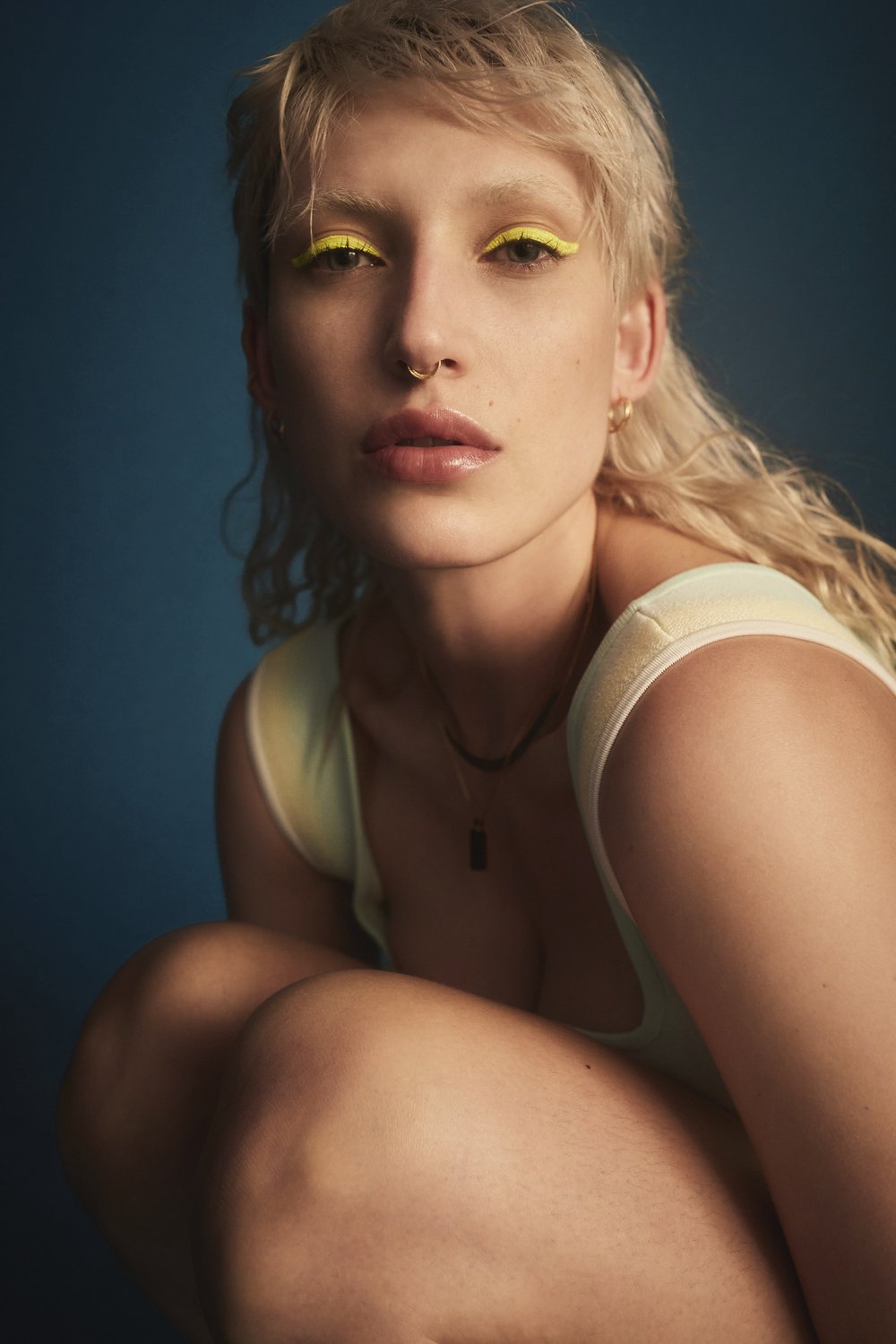 Close-up portrait of a fair-skinned woman with bleached blonde hair and yellow eyeliner posing against a dark blue background.