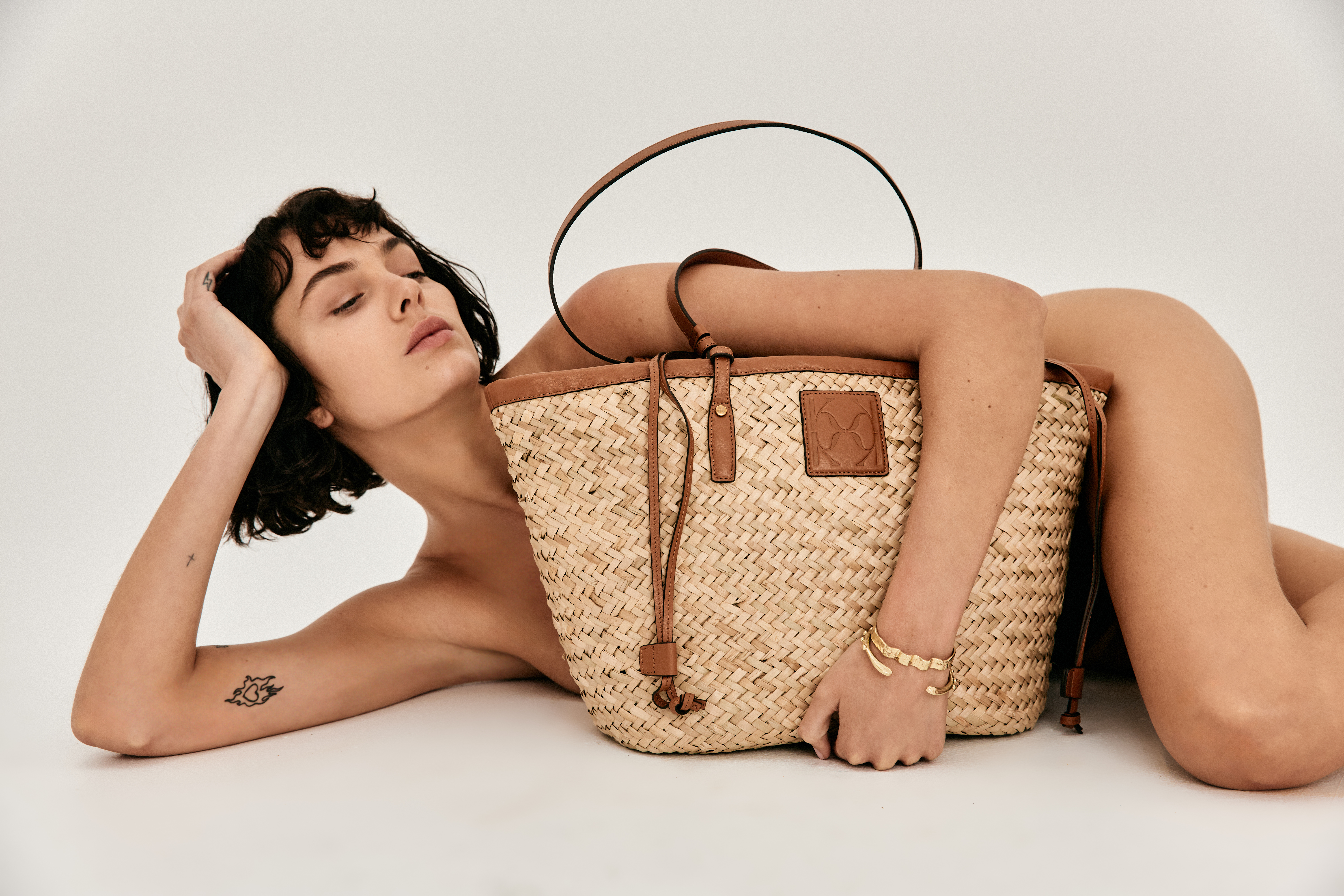 Woman poses with straw tote bag with leather trim in a studio setting. The bag is the primary focus in a light-colored scene.