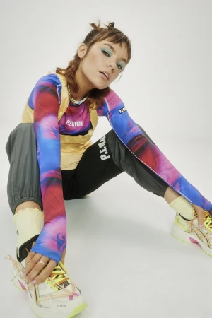 Young woman with fair skin, wearing a tie-dye-style long-sleeve top, grey track pants, and yellow sneakers, poses in a studio.