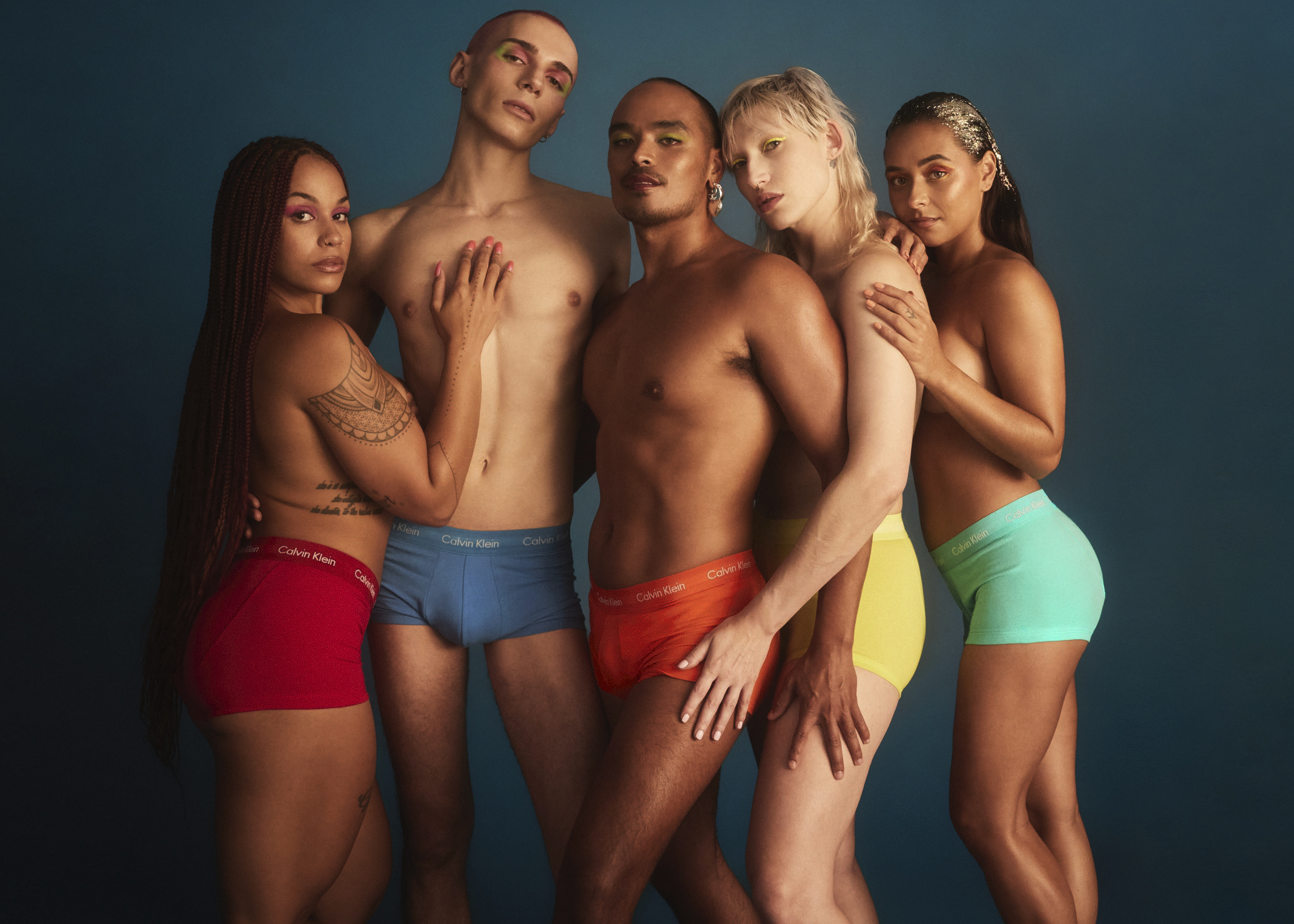 Five diverse people pose together in a studio wearing red, blue, orange, yellow, and green briefs with a teal background.