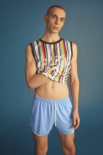 A person with short pink hair poses in a rainbow-striped crop top and blue shorts against a blue backdrop in a studio shot.