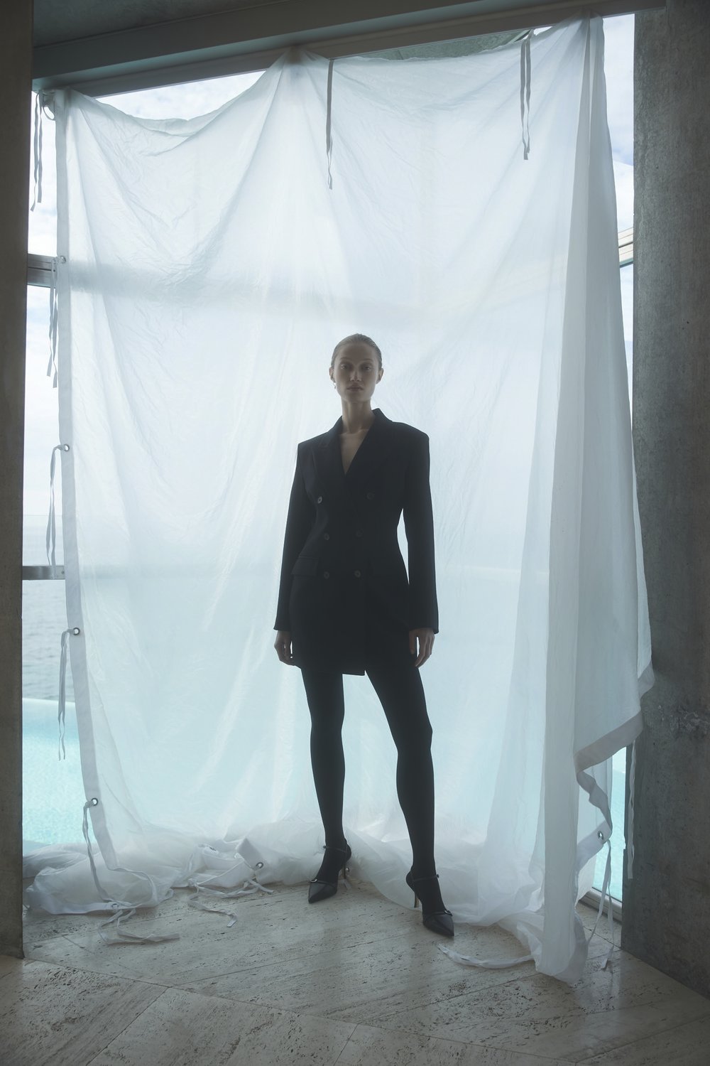 Woman poses in a black blazer dress in front of a sheer white curtain, indoor, against a window view of a pool.