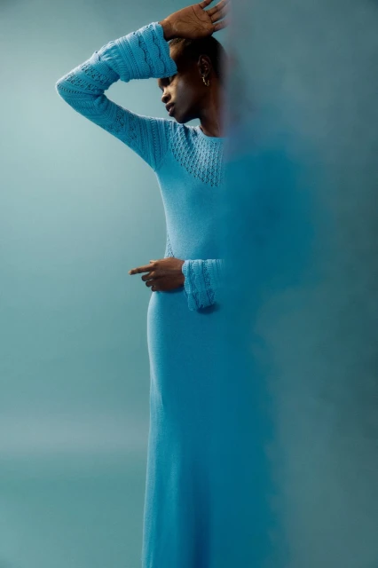 Black fashion model poses in a blue dress in a studio setting. The image features soft lighting, with a focus on the model's silhouette.
