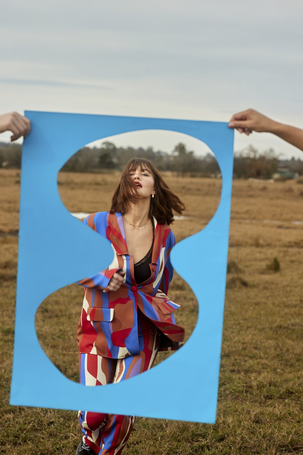 Woman in colorful suit poses behind blue abstract frame in an outdoor field setting with overcast sky and sparse trees in the distance.