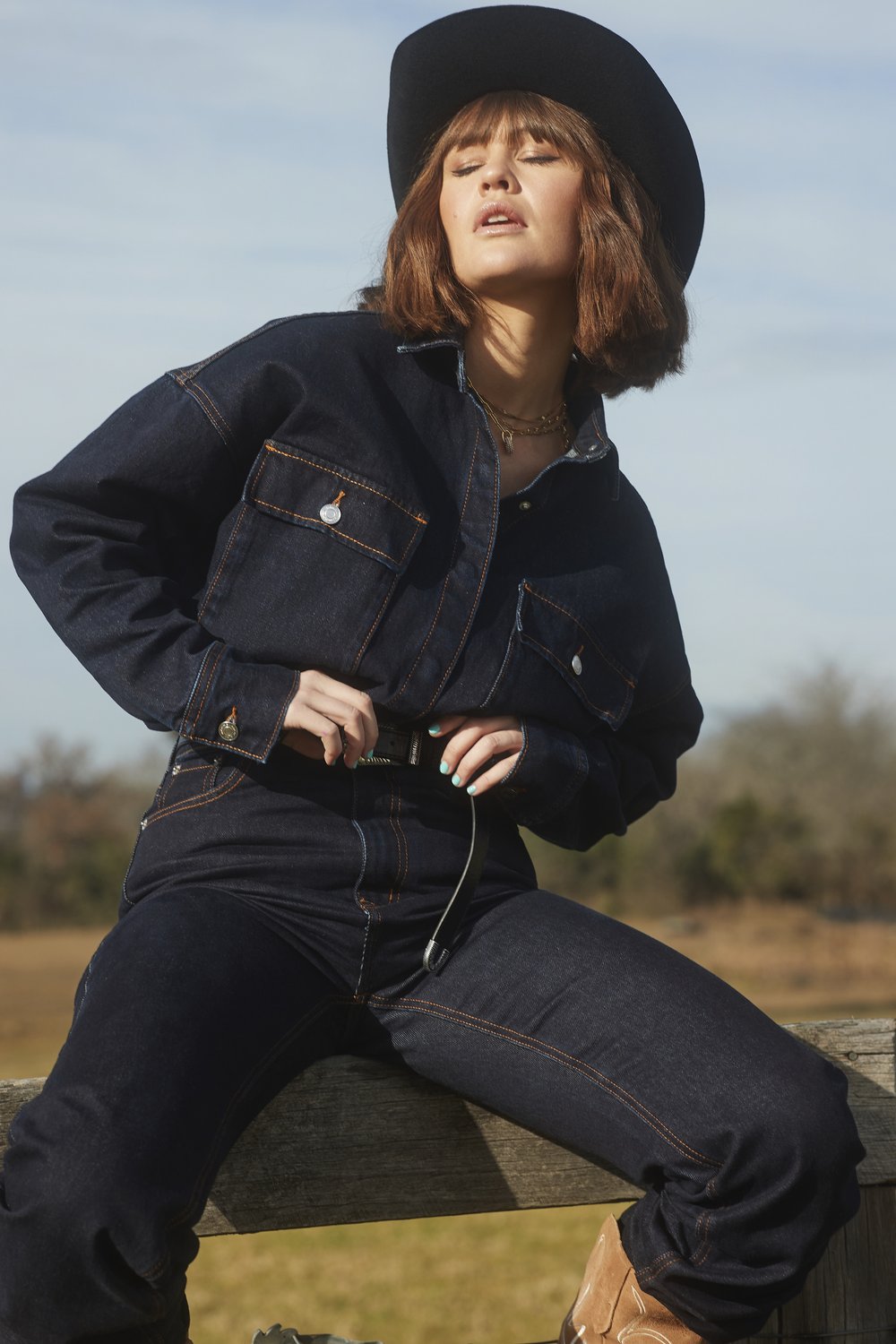 A woman in a dark denim jacket and jeans with a cowboy hat sits on a wooden fence, bathed in natural light, set against a rural backdrop.