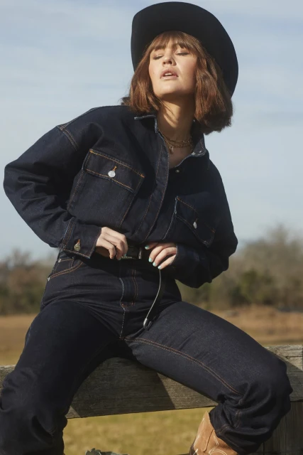 A woman in a dark denim jacket and jeans with a cowboy hat sits on a wooden fence, bathed in natural light, set against a rural backdrop.