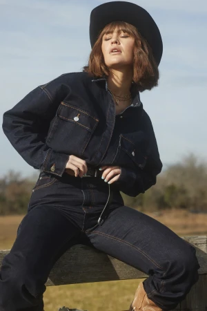 A woman in a dark denim jacket and jeans with a cowboy hat sits on a wooden fence, bathed in natural light, set against a rural backdrop.