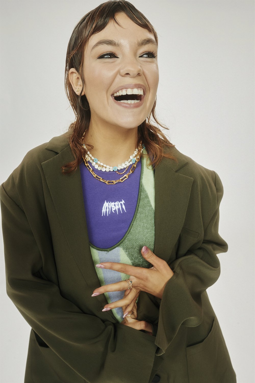 Smiling woman in a green jacket and stylish necklaces, posing in front of a plain white background in a studio setting. Beauty shot.