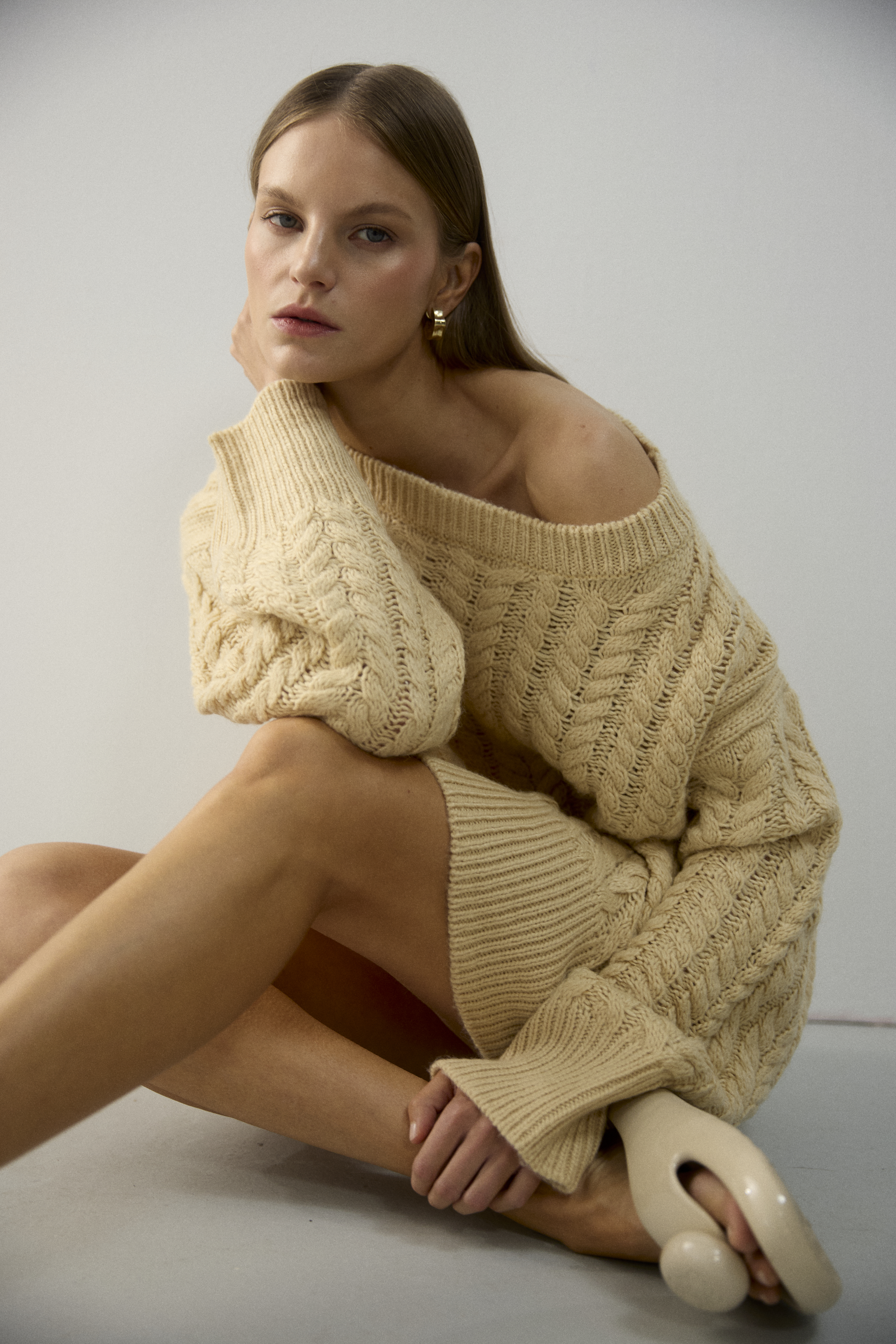 A blonde-haired woman in an off-the-shoulder cable knit sweater dress sits on the floor in a minimalist studio with modern sandals.