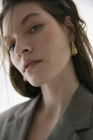Close-up shot of a young adult woman with fair skin, wearing a grey pinstripe blazer and golden earrings, posing against a white background.