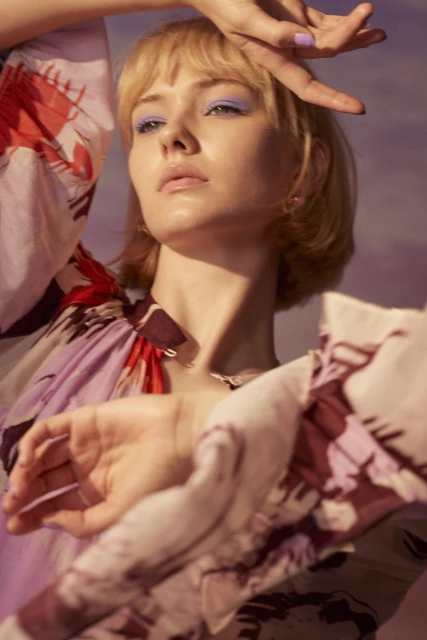 Blonde female model with floral blouse, hands raised in a studio setting, displaying light purple eyeshadow and matching nail polish, artistic pose.