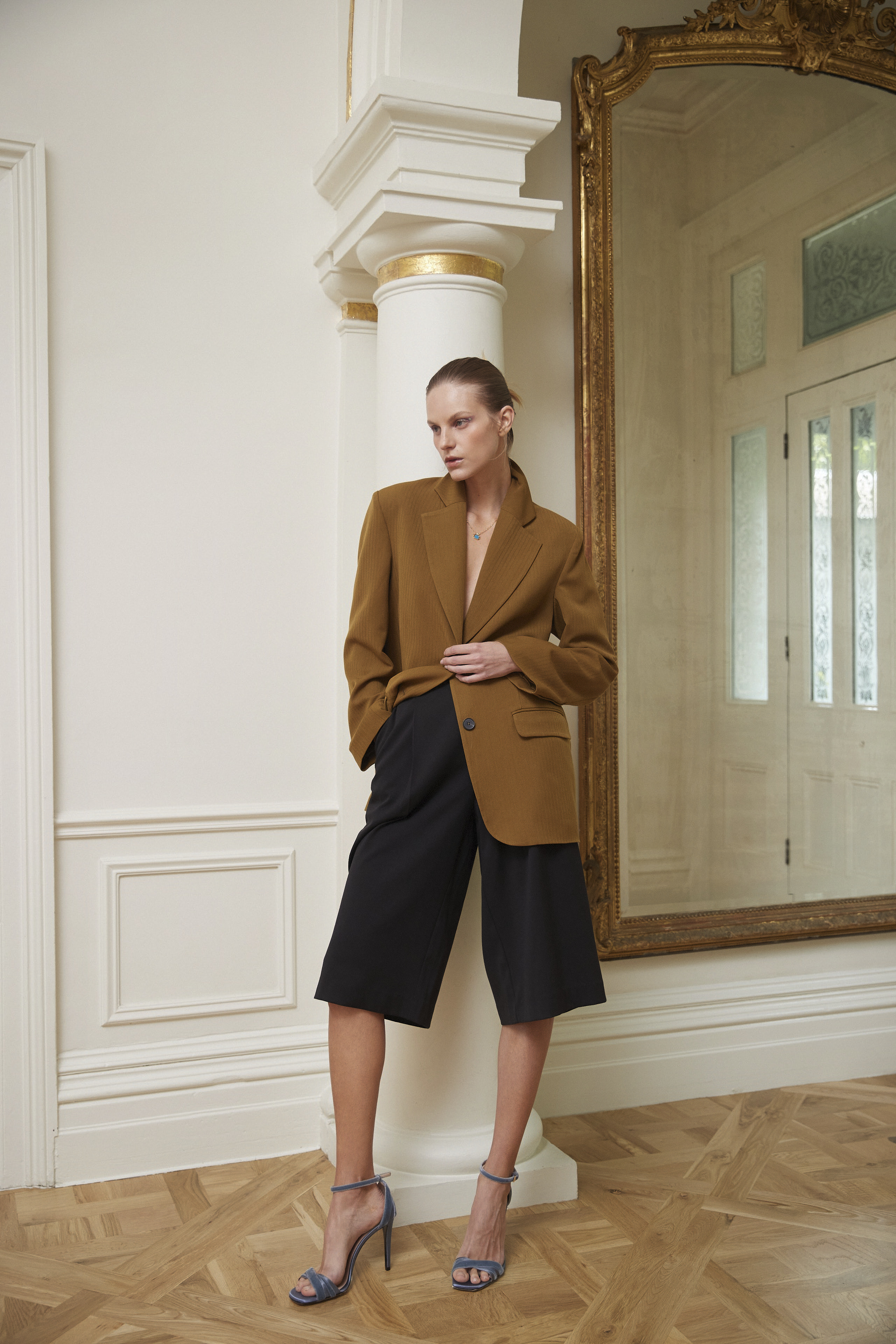 A woman poses in a brown blazer and black culottes, standing in front of a white column and ornate mirror, showcasing fashion in an elegant setting.