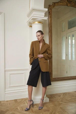 A woman poses in a brown blazer and black culottes, standing in front of a white column and ornate mirror, showcasing fashion in an elegant setting.