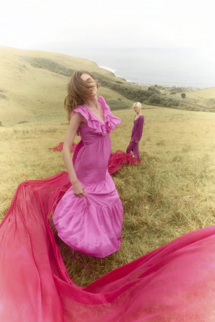 Two women in pink dresses stand in a grassy field overlooking the ocean and hills. One woman has long flowing fabric trailing behind her.