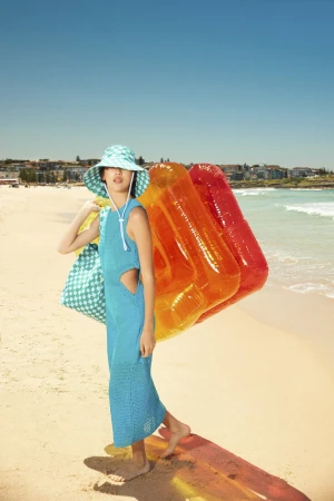 Woman in blue dress and hat on beach, holding orange inflatable raft, blue sky background. Vacation fashion editorial shot at ocean.