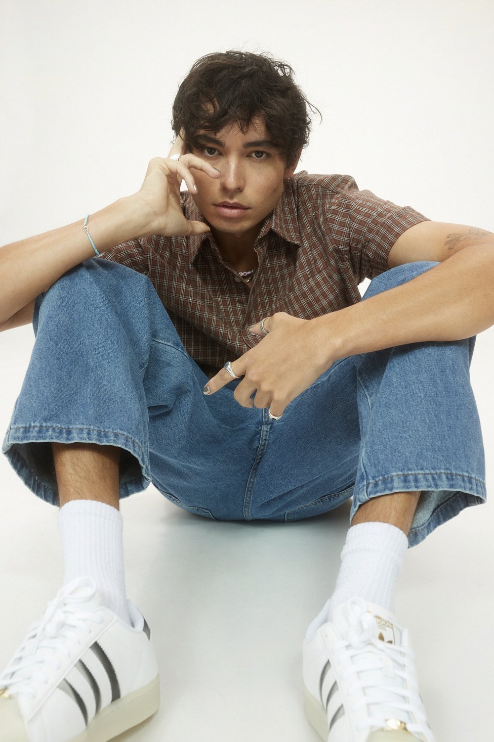 Portrait of a young adult male in a brown checkered shirt and blue jeans, sitting on the floor in a photo studio.
