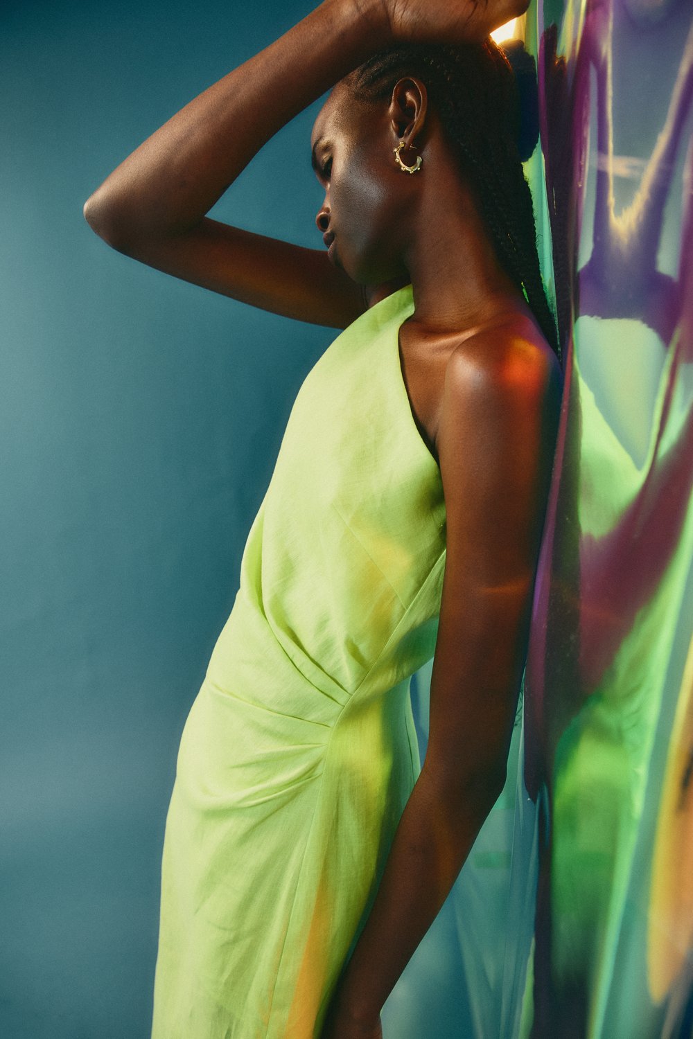 Woman in a light green dress leans against a colorful reflective surface in a studio setting with soft lighting, showcasing fashion.