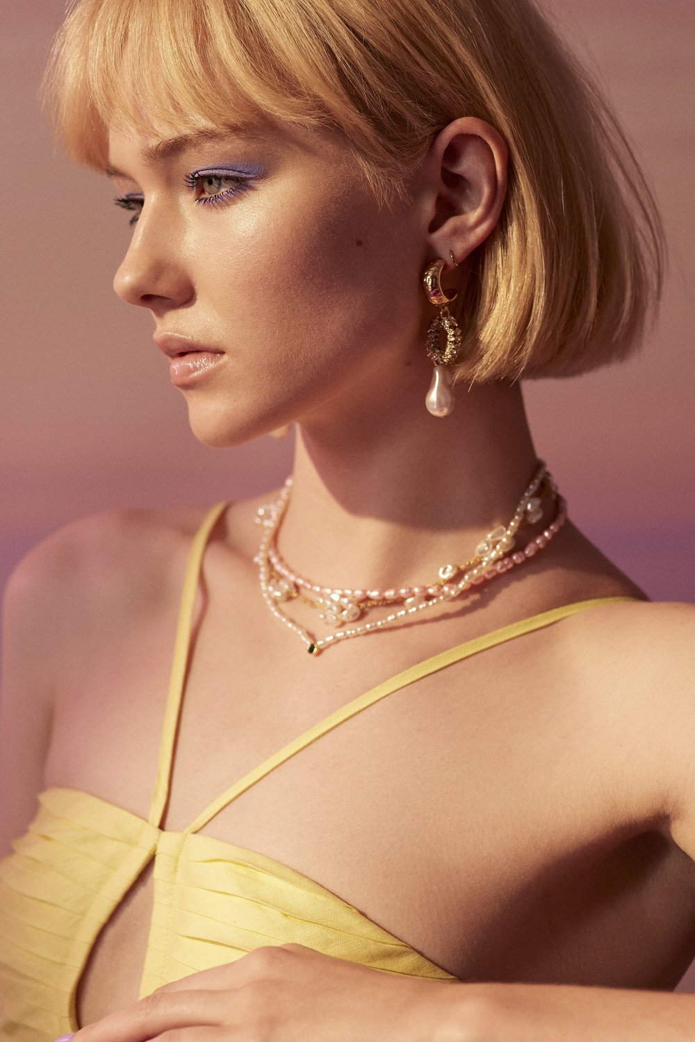 Close-up profile of a fair-skinned woman with blonde hair, wearing a yellow dress and layered pearl necklaces against a soft gradient background.