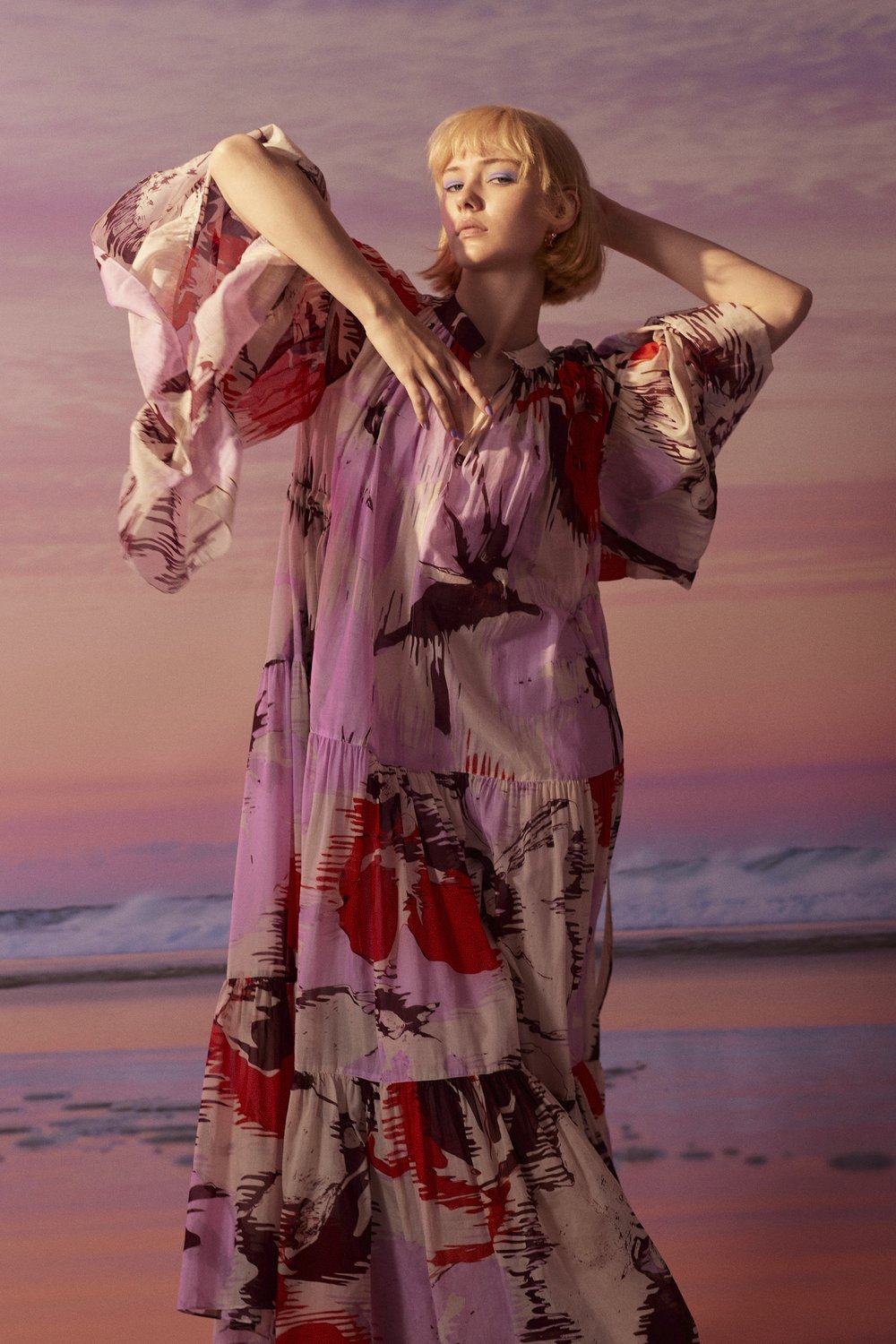 Young woman in a flowing floral dress posing against a softly blurred seascape backdrop, fashion shot, warm lighting, soft focus.