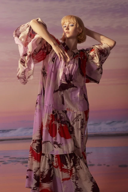 Young woman in a flowing floral dress posing against a softly blurred seascape backdrop, fashion shot, warm lighting, soft focus.