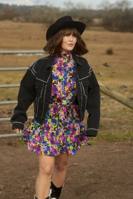 A woman in a black cowboy hat, jacket with studs, and floral dress poses in a rural field with a blurred background.