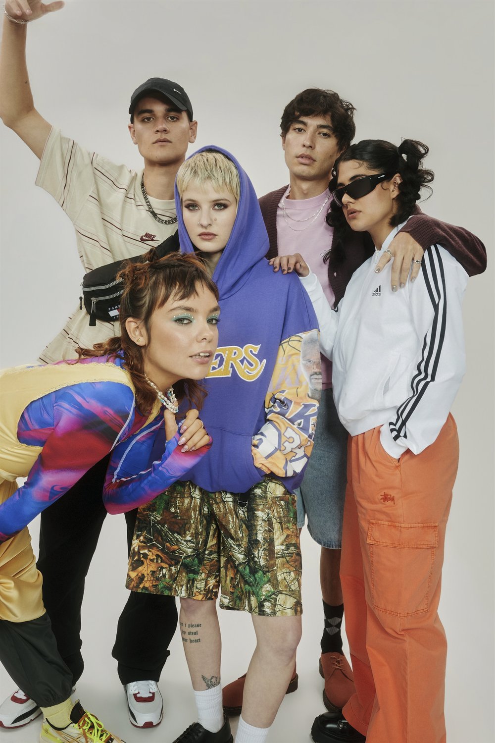 Group of five young adults in streetwear fashion posing in a photo studio with a white backdrop, showcasing diverse styles and brands.