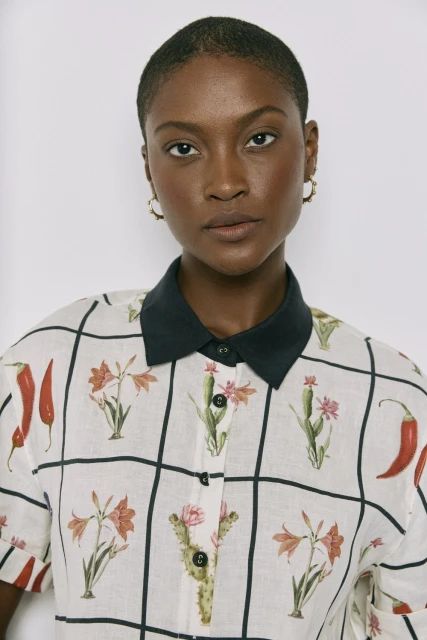 Portrait of a Black woman wearing a botanical-print collared shirt in front of a neutral background, facing forward with a serious expression.