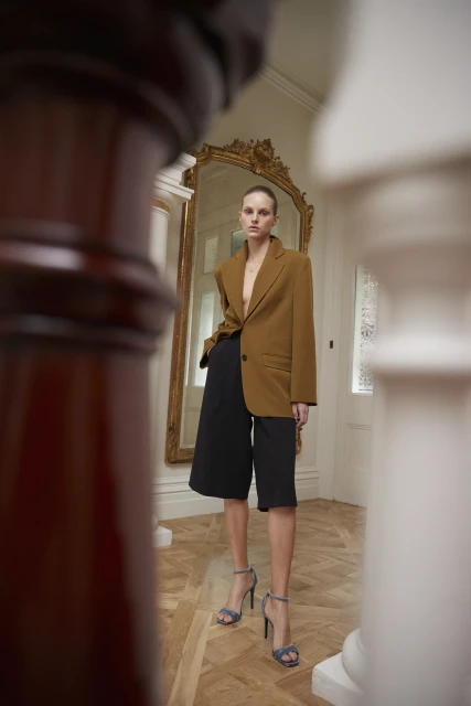 A stylish woman in an oversized brown blazer and black culottes strikes a pose indoors near a gilded mirror in a fashion shot.