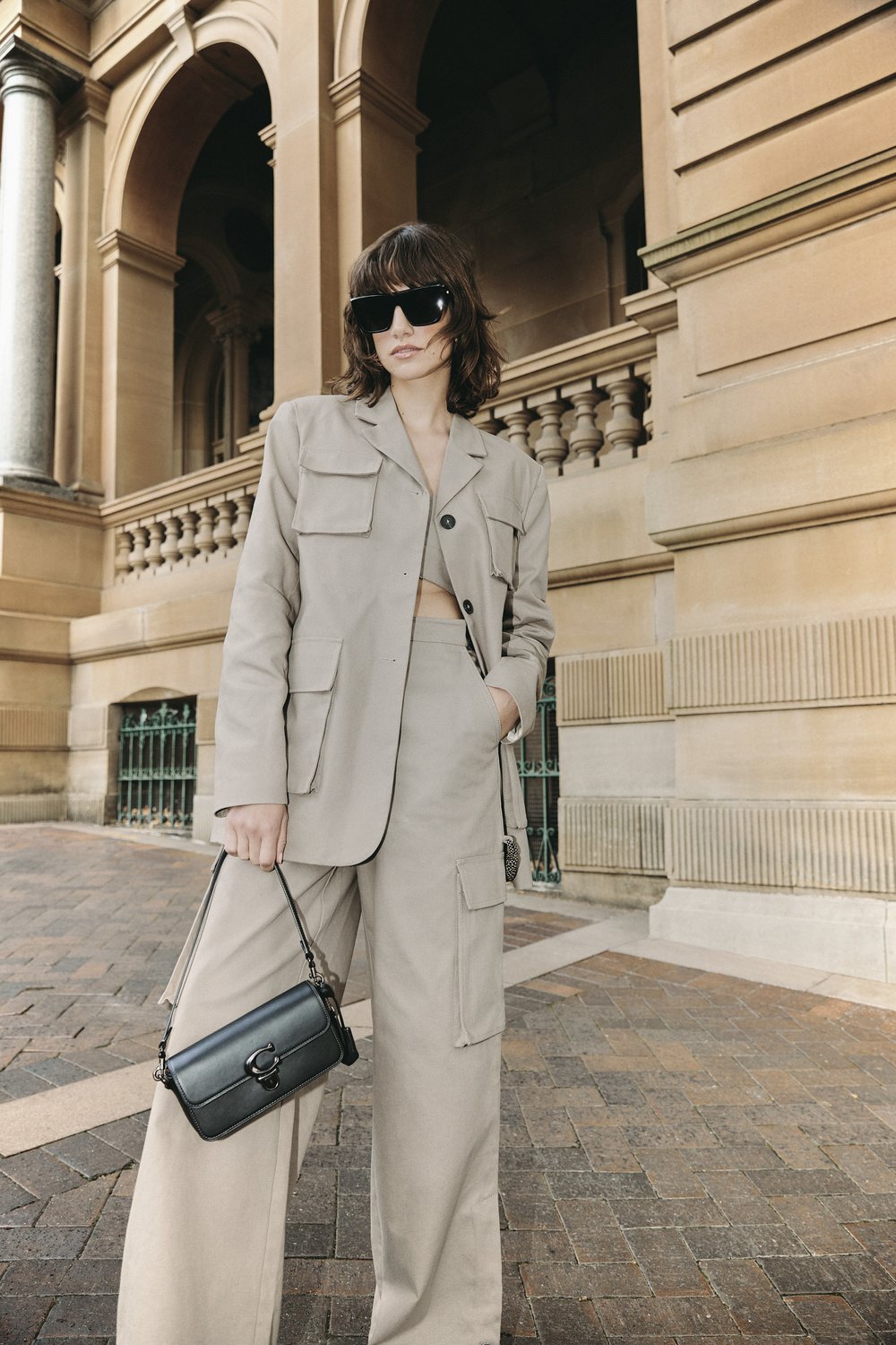 A woman in a tan blazer and wide-leg pants poses outdoors in front of a building with arches, carrying a black Coach purse.